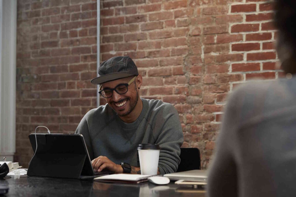 A man in glasses and a cap laughing while typing on a tablet at a table