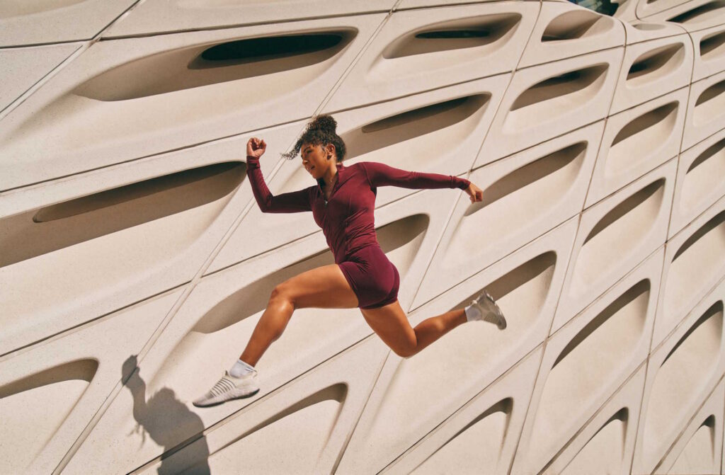 Female Athlete Makes A Running Jump Near The Geometrical Facade Of A Building