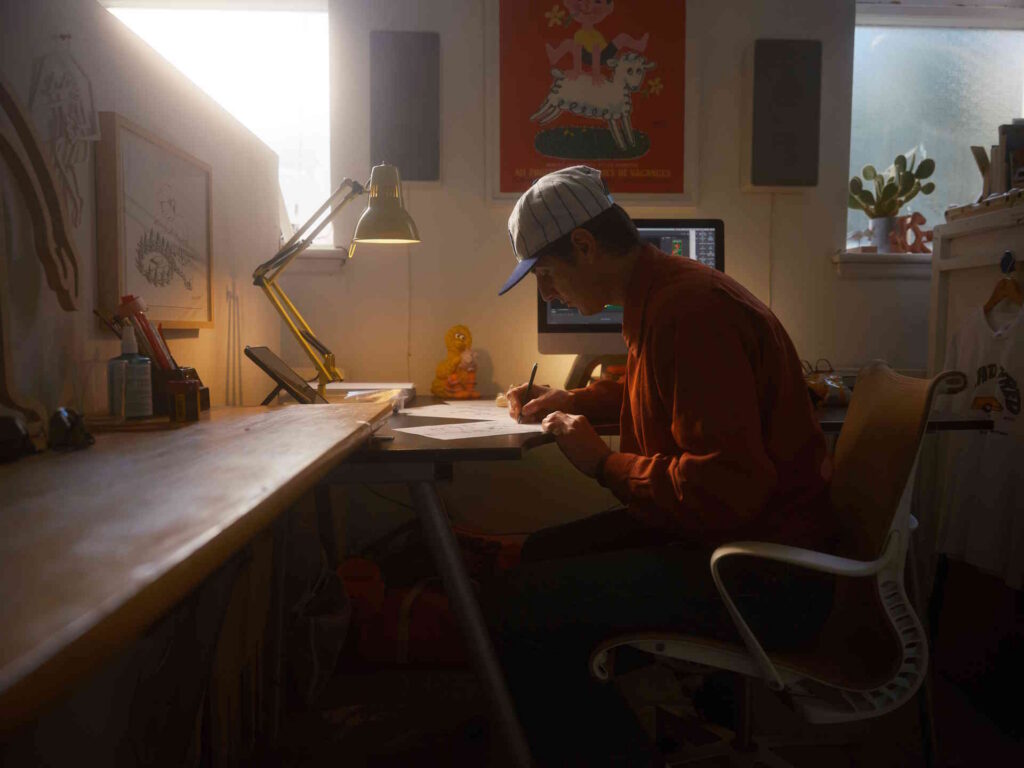 A man in a baseball cap sketching by hand at a lit desk in a moody, warm-toned studio, computer monitor and childhood-toy figurines in the background