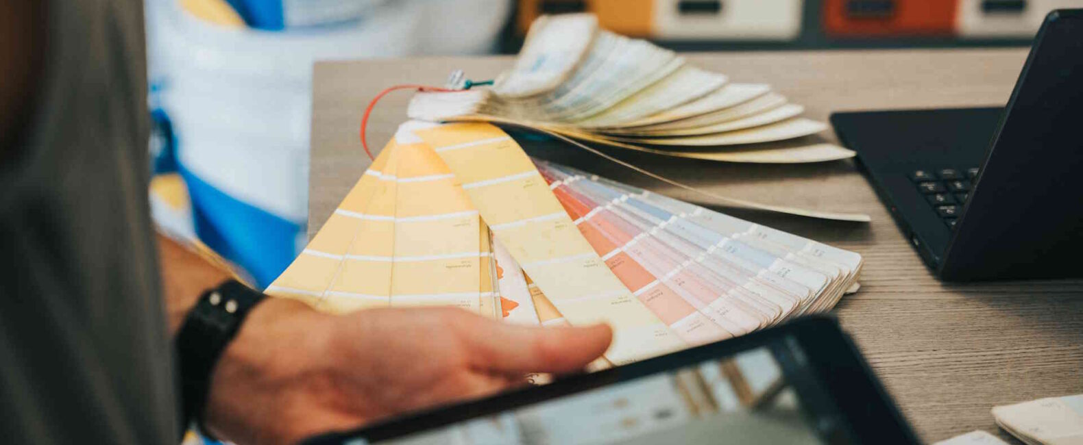 A man's hands flipping through paint color swatches while holding a tablet displaying a similar image
