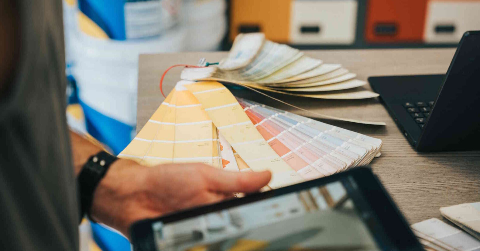 A man's hands flipping through paint color swatches while holding a tablet displaying a similar image