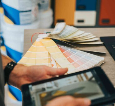 A man's hands flipping through paint color swatches while holding a tablet displaying a similar image