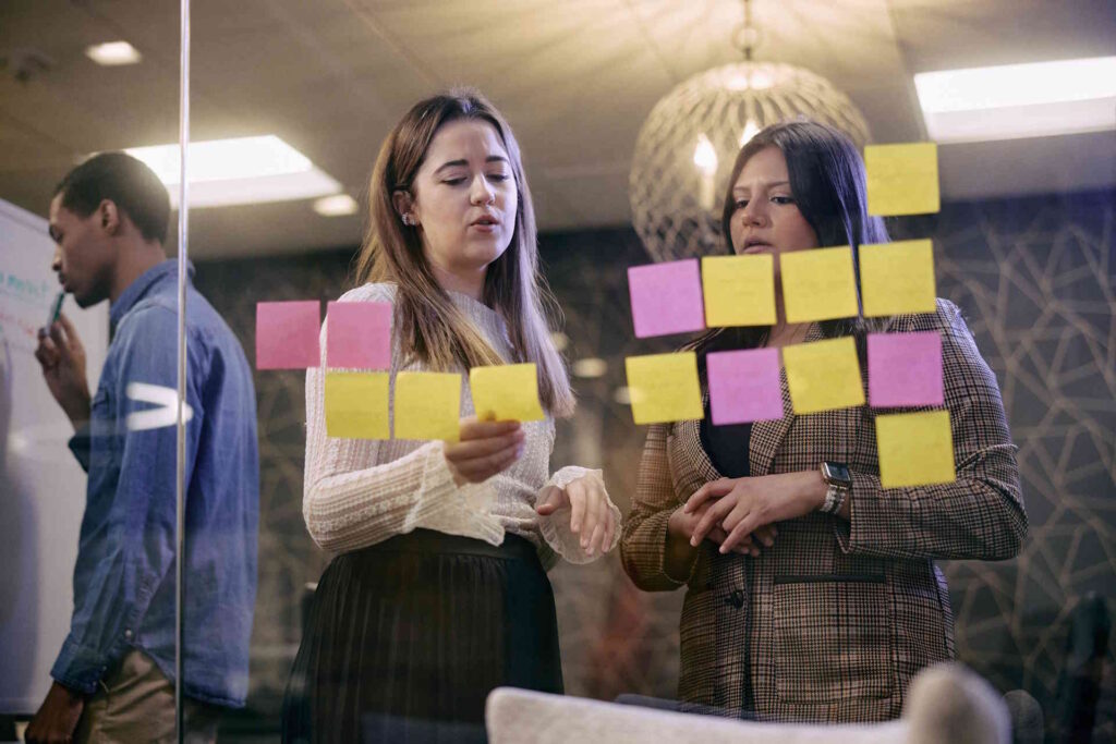 Two women through a glass wall arranging pink and yellow sticky notes, with a man writing on a whiteboard in the background