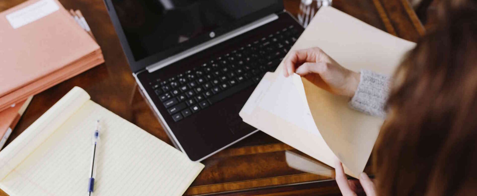 Overhead shot of a woman at a wood desk flipping through a folder beside an open laptop and notepad