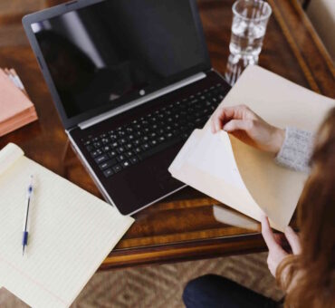 Overhead shot of a woman at a wood desk flipping through a folder beside an open laptop and notepad