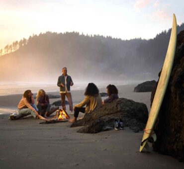 small group gathered around a beach bonfire at dusk, with a wetsuit and surfboard nearby