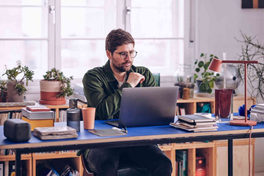 A man at a cluttered, plant-filled desk studying his laptop screen with a focused, unsmiling expression