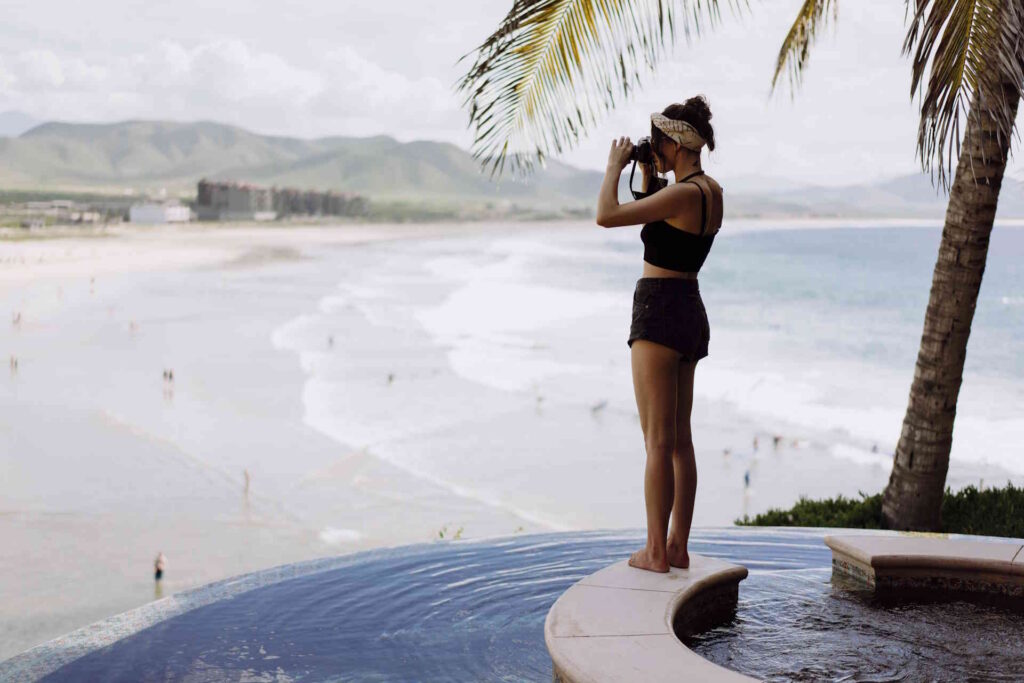 A woman standing at the edge of an infinity pool overlooking a beach, photographing the view with a real camera.
