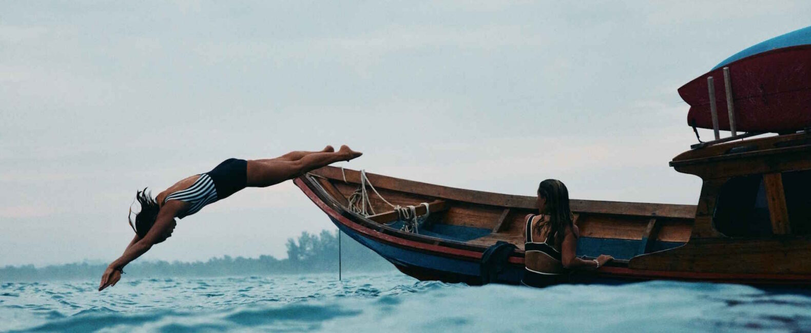 A Woman In A Striped Swimsuit Dives Gracefully From A Wooden Boat
